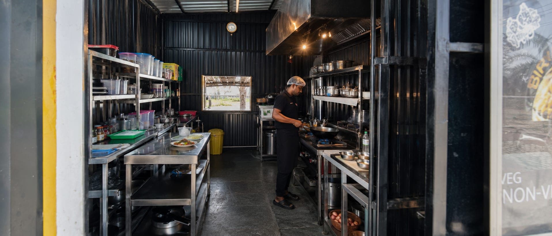 The open kitchen seen from the service window: a cook working a wok over the range, stainless steel counters and shelves of stock along both walls.