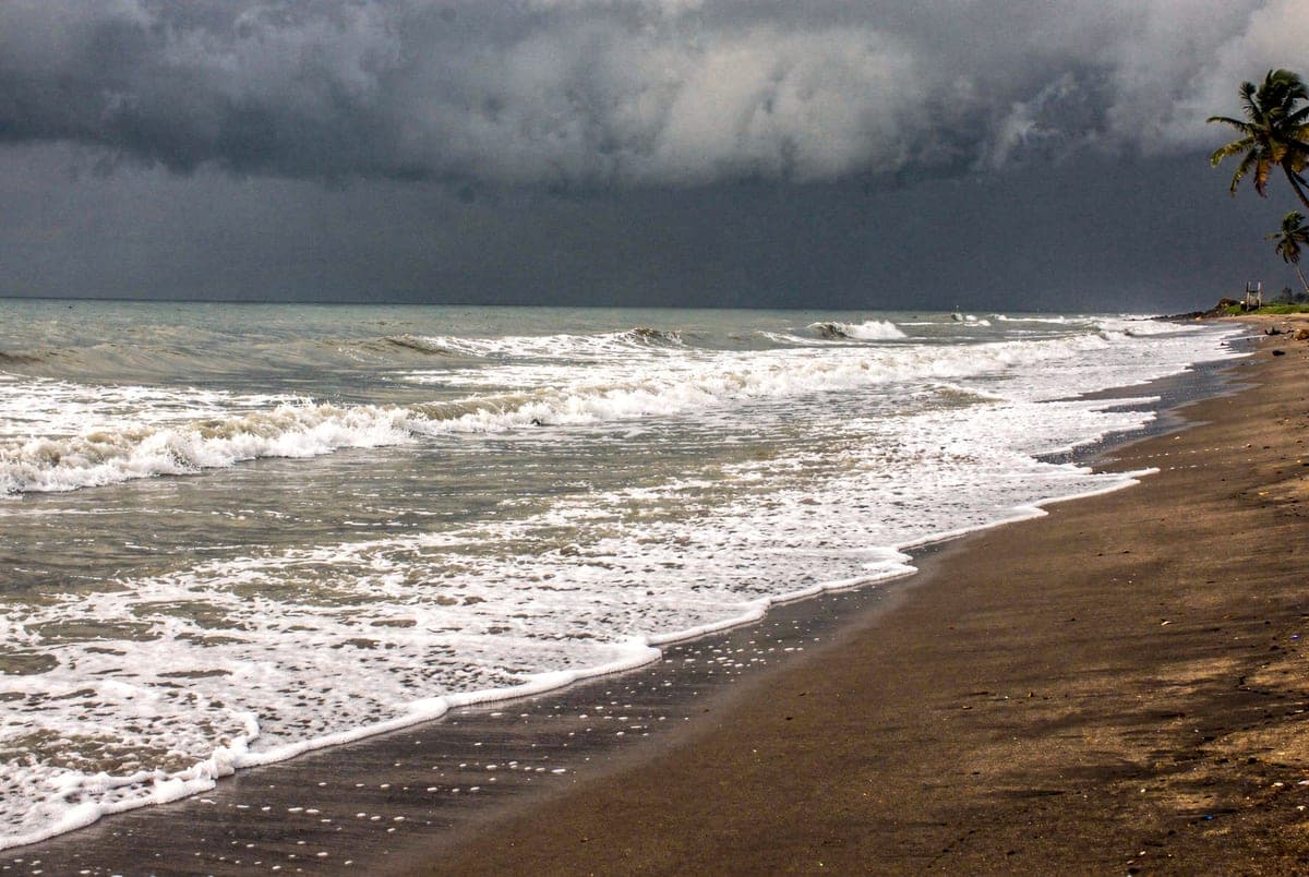 Grey-green surf breaking on dark wet sand under a heavy bank of monsoon cloud.